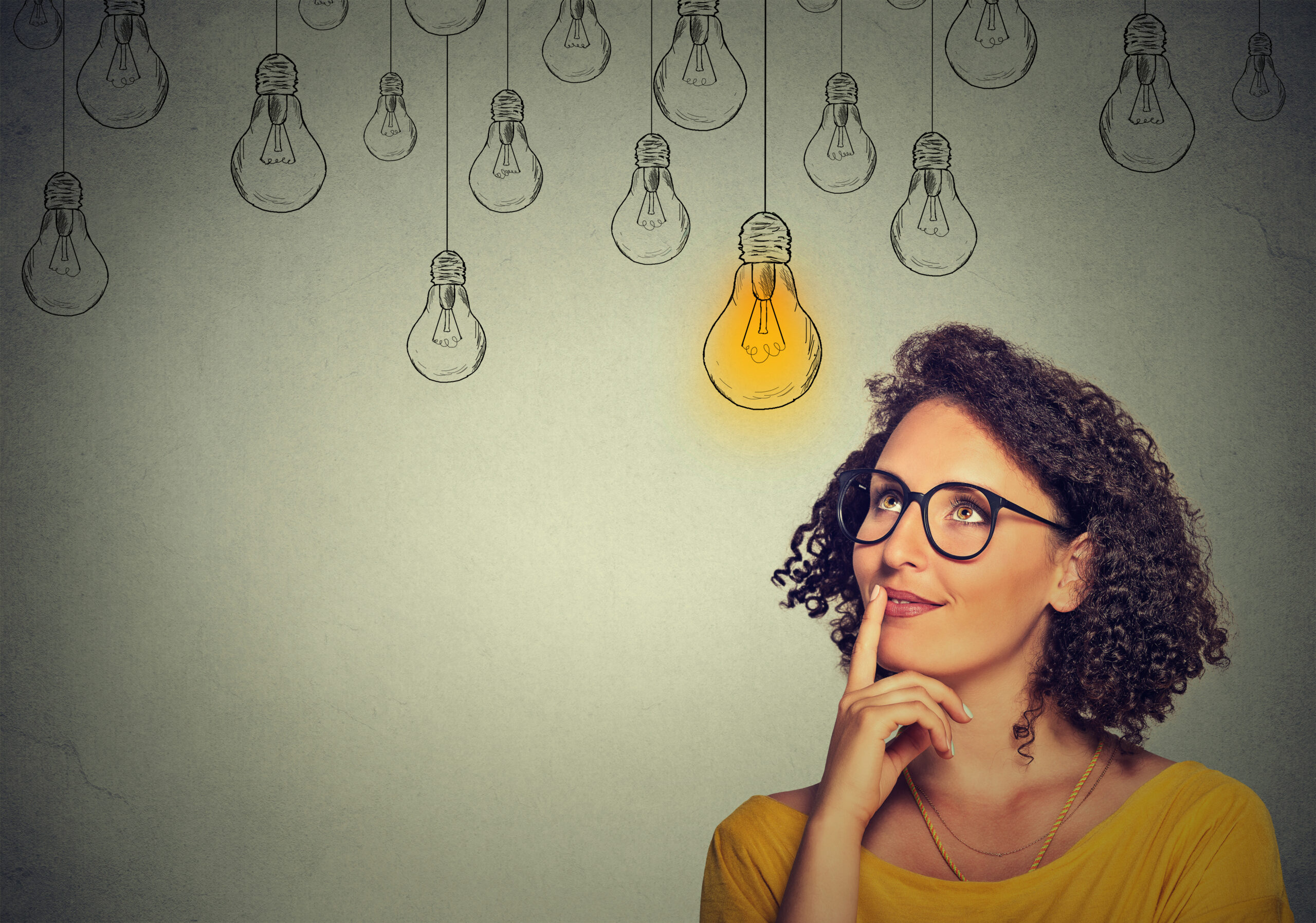 Portrait thinking woman in glasses looking up with light idea bulb above head isolated on gray wall background
