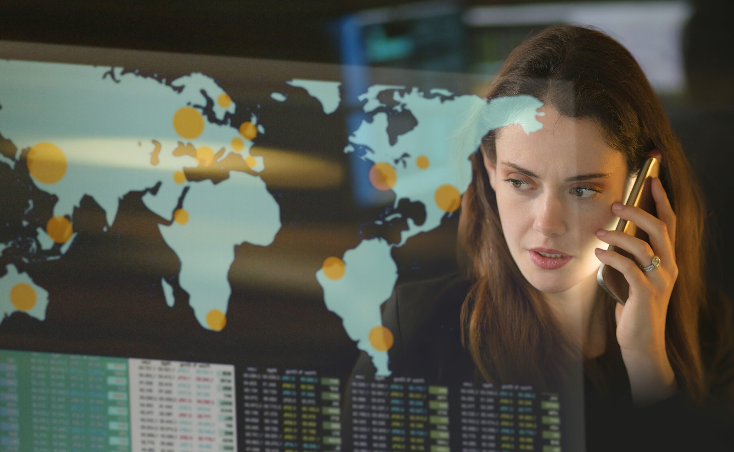 Stock image of a good looking woman talking on the phone in a dark office. She’s at a see-through screen depicting the world with numbers and data.