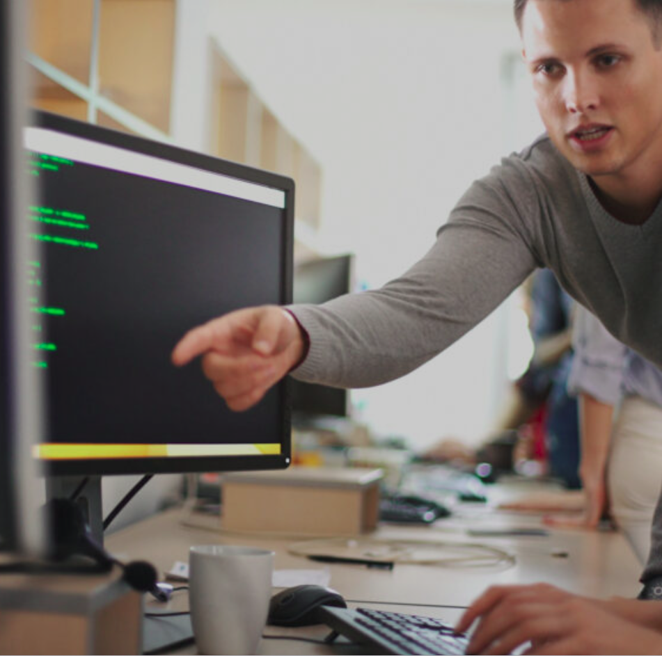 a young man pointing at a monitor screen, whilst talking to a fellow worker